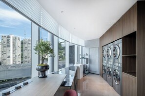 Une chambre d’hôtel moderne avec de grandes fenêtres, une plante en pot, un lave-linge et une vue sur de hauts immeubles.