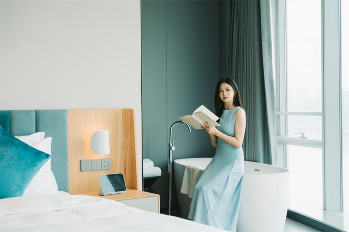 A modern hotel room with a bed, a bedside table with a lamp, a large window with curtains, and a woman reading a book.