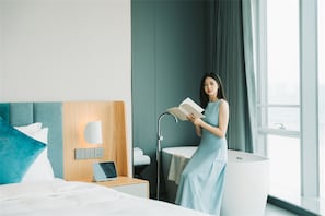 A modern hotel room with a bed, a bedside table with a lamp, a large window with curtains, and a woman reading a book. - Pagoda Hotel Chongqing Near By Liziba Hongya Cave (Chongqing)