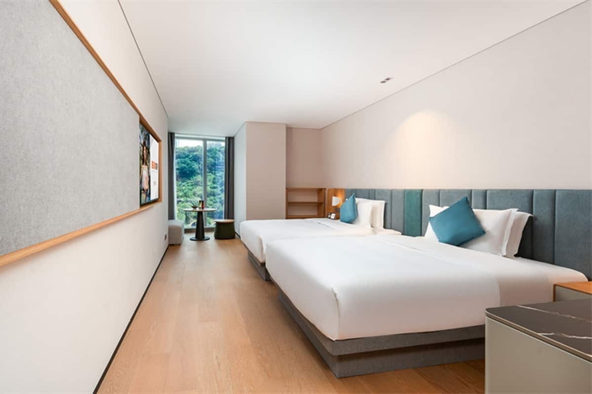 A modern hotel room with a large bed, a desk, and a view of greenery outside.