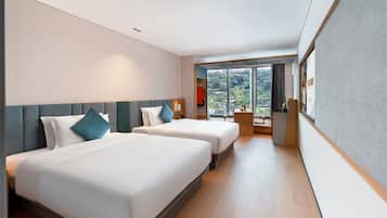 A hotel room with two beds, a small desk, a window with a view of greenery, and a wardrobe.