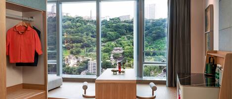 A modern hotel room with a large window offering a view of a lush green hillside. There is a wooden bench with a small table and decorative items in front of the window.