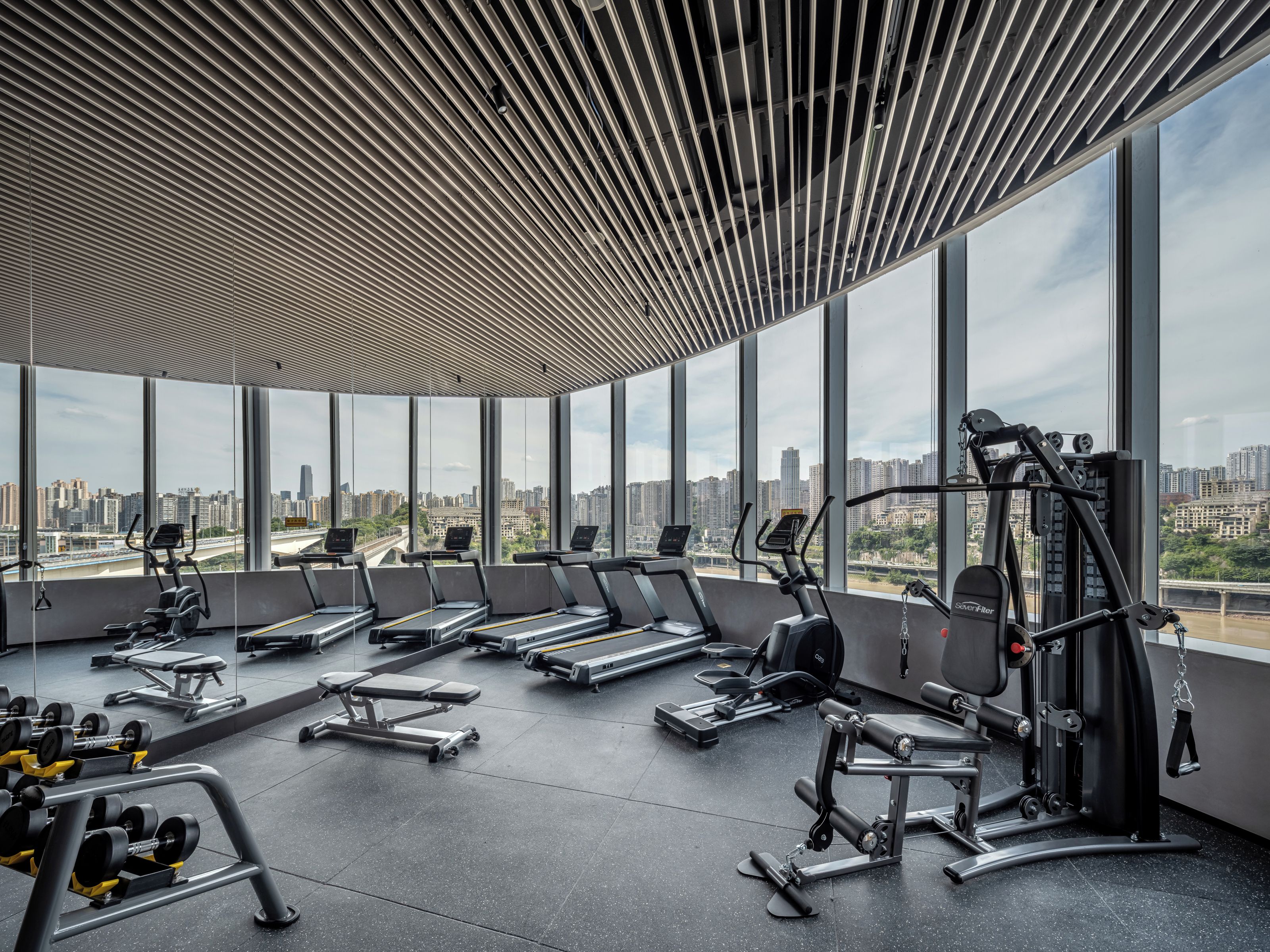 Un gimnasio moderno con cintas de correr, máquinas de pesas y bancos, que cuenta con ventanas amplias y vistas a la ciudad.