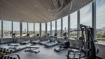 Un gimnasio moderno con cintas de correr, máquinas de pesas y bancos, que cuenta con ventanas amplias y vistas a la ciudad.