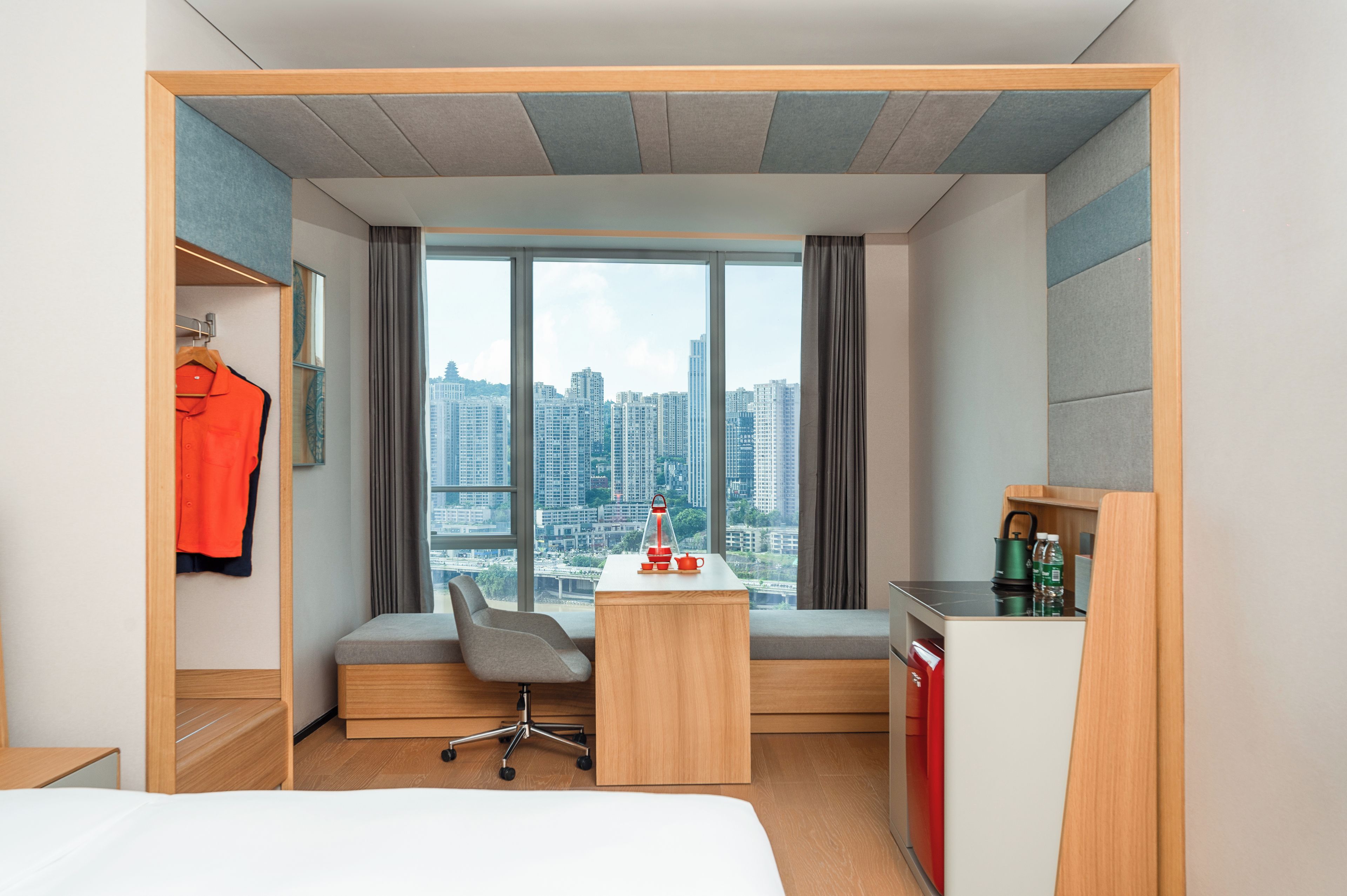 A hotel room with a bed, a desk, a chair, a wardrobe, a window with a city view, and a small kitchenette.
