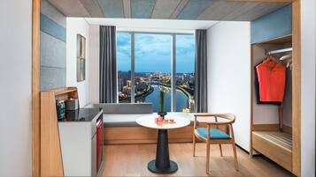 Habitación de hotel moderna con un ventanal grande, una mesa redonda, un sofá, una silla, una pequeña cocina y un armario.