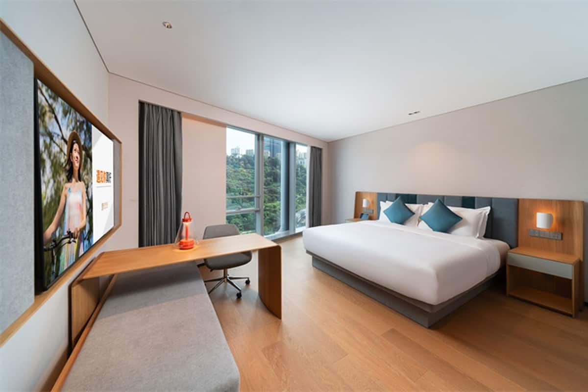 A modern hotel room with a large bed, a desk, a chair, and a view of greenery outside.