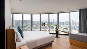 A modern hotel room with a large bed, a small table, and a view of a cityscape through large windows.