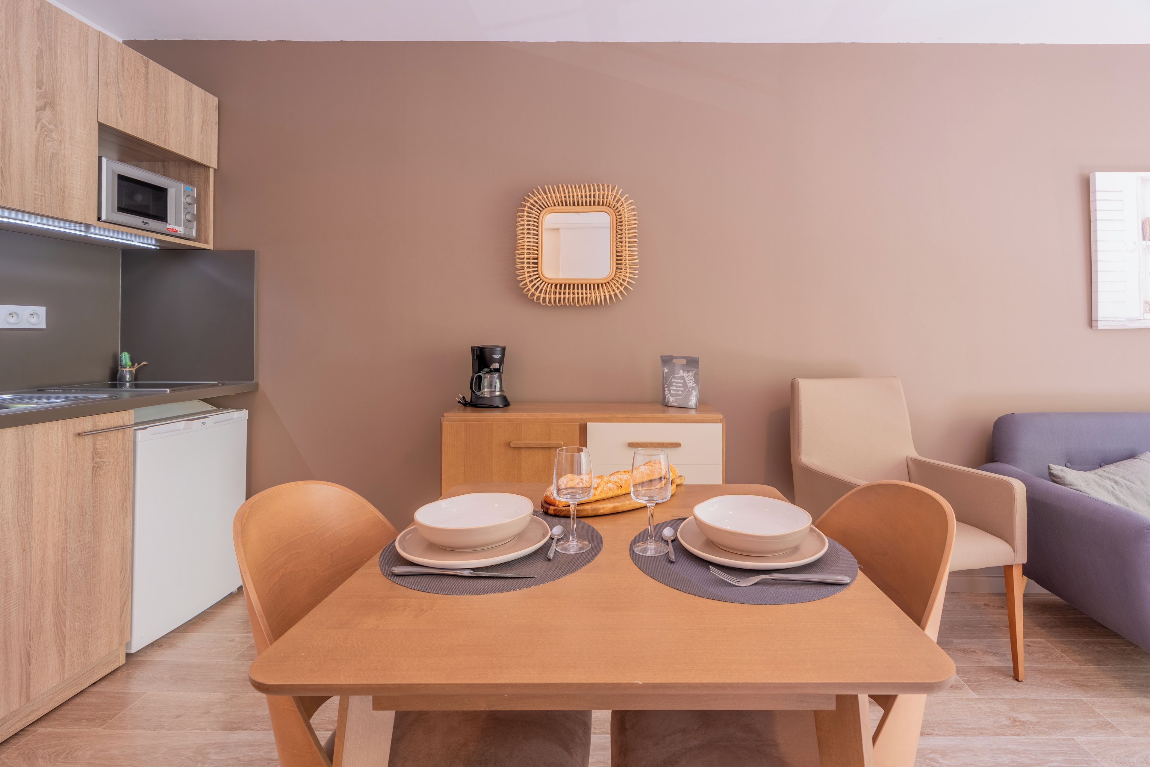 Une cuisine moderne équipée d’un four à micro-ondes, d’une machine à café et d’un petit réfrigérateur. On y trouve une table à manger, mise avec des assiettes, des verres et un pain. Un miroir en osier est accroché au mur, au-dessus de la table.