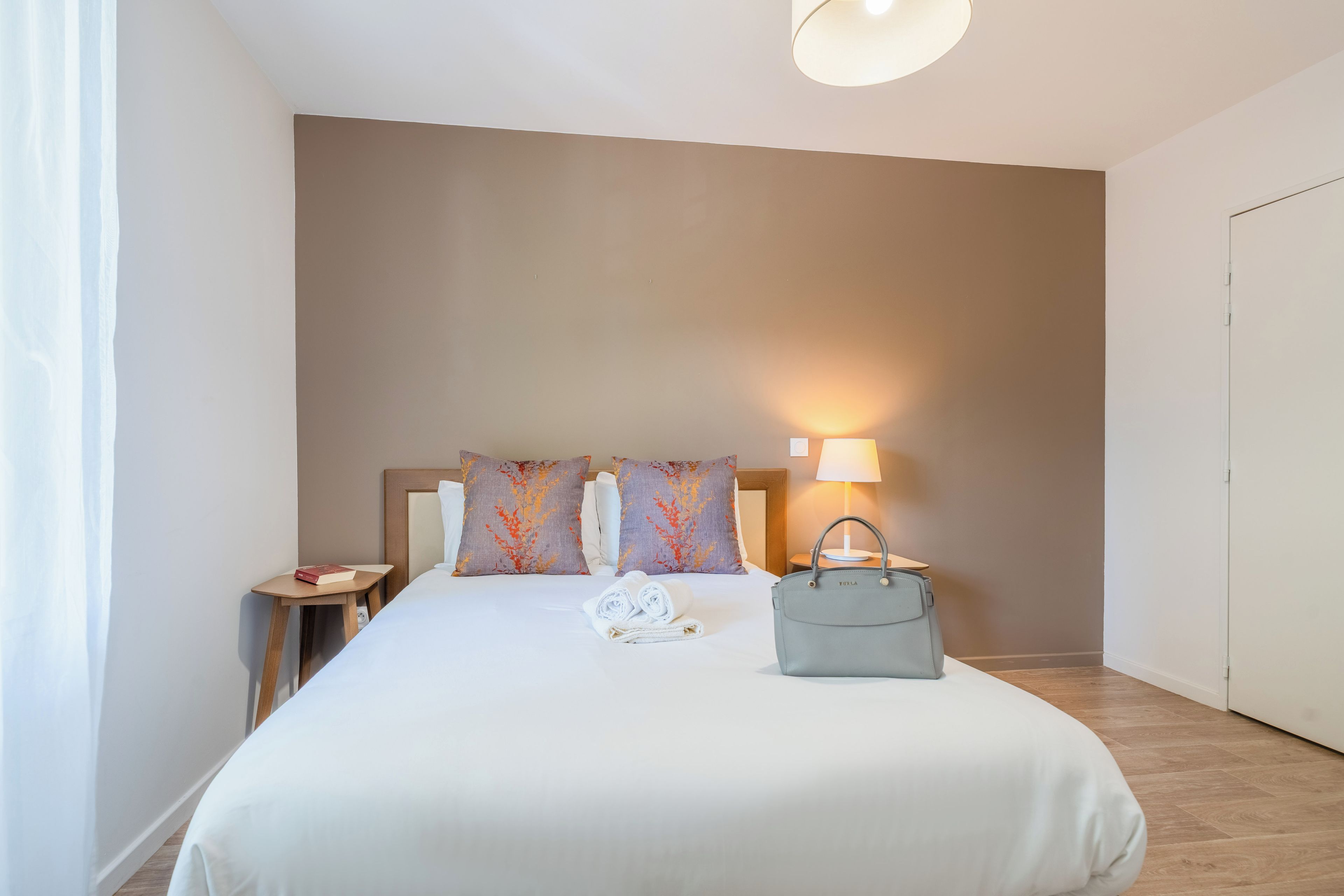 A neatly made bed with pillows, a bedside lamp, a small table with a book, and a handbag.
