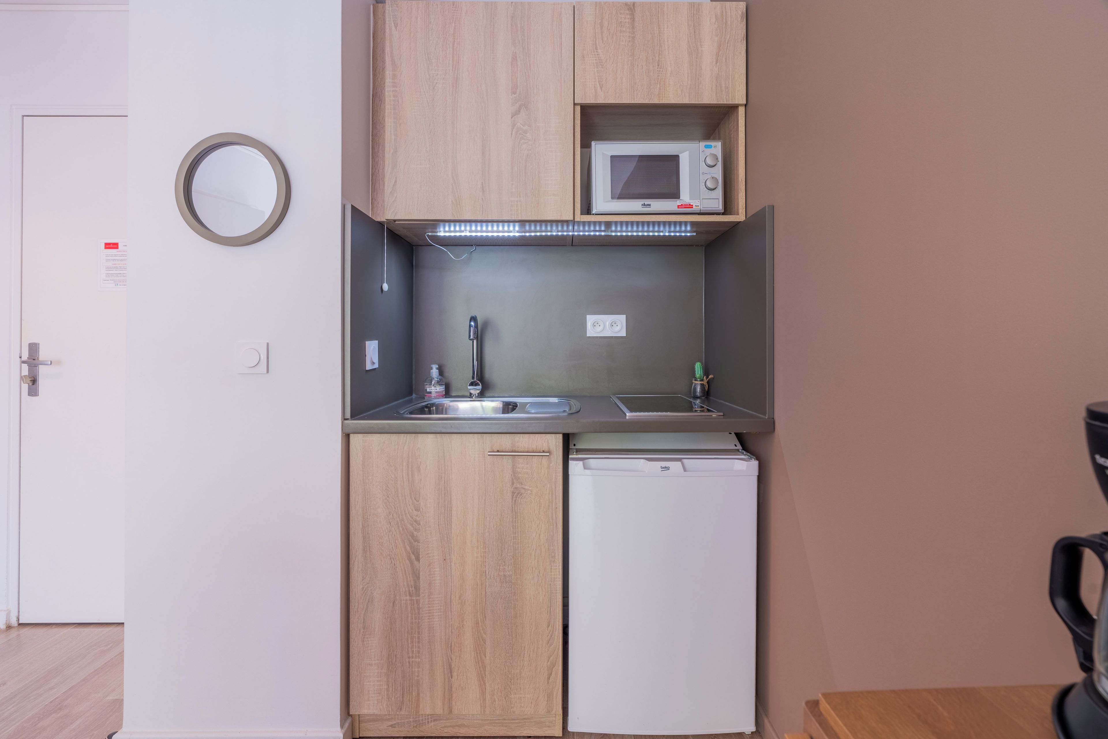 A compact kitchenette with a microwave, sink, and dishwasher, wooden cabinets, and a round mirror.