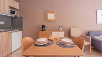 Una cocina moderna con microondas, cafetera y un refrigerador pequeño. Hay una mesa de comedor con platos, vasos y una hogaza de pan. Un espejo de mimbre cuelga en la pared sobre la mesa.