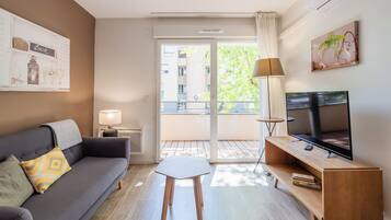 Una sala moderna con sofá, una mesita, televisor y un balcón con vistas a edificios y árboles.