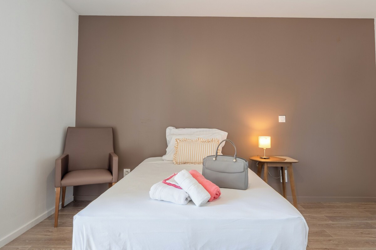 A hotel room with a bed, a chair, a bedside table with a lamp, and a wall-mounted artwork.