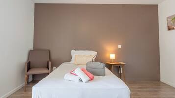 A hotel room with a bed, a chair, a bedside table with a lamp, and a wall-mounted artwork.