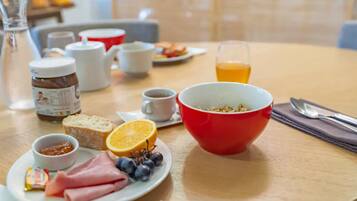 Un desayuno con un tazón rojo, platos de jamón, pan y fruta, un vaso de zumo de naranja y una taza de café.