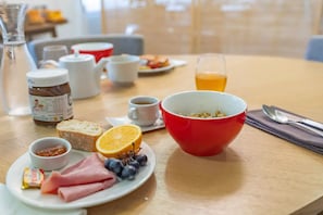 A breakfast setting with a red bowl, plates with ham, bread, and fruit, a glass of orange juice, and a coffee cup. - Quality Aparthotel Lyon L'isle-dabeau (L'Isle-d'Abeau)