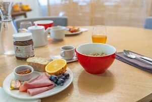Un desayuno con un tazón rojo, platos de jamón, pan y fruta, un vaso de zumo de naranja y una taza de café.