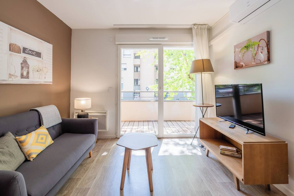 Uma sala de estar moderna com um sofá, uma mesinha, uma televisão e uma varanda com vista para edifícios e árvores.