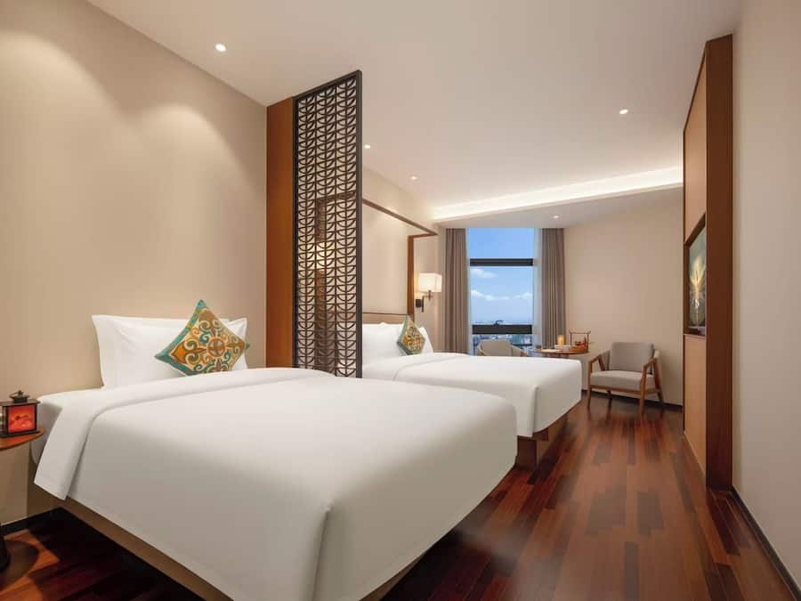 Habitación de hotel con dos camas, una mesita de noche, una silla y vista a la ciudad.