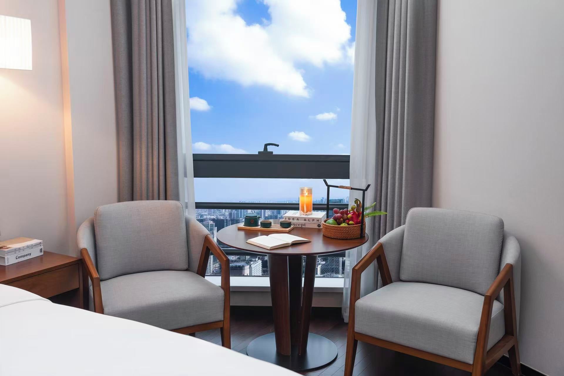 Habitación de hotel con dos sillones grises tapizados, una mesa redonda de madera con una vela, un libro y una cesta de frutas.