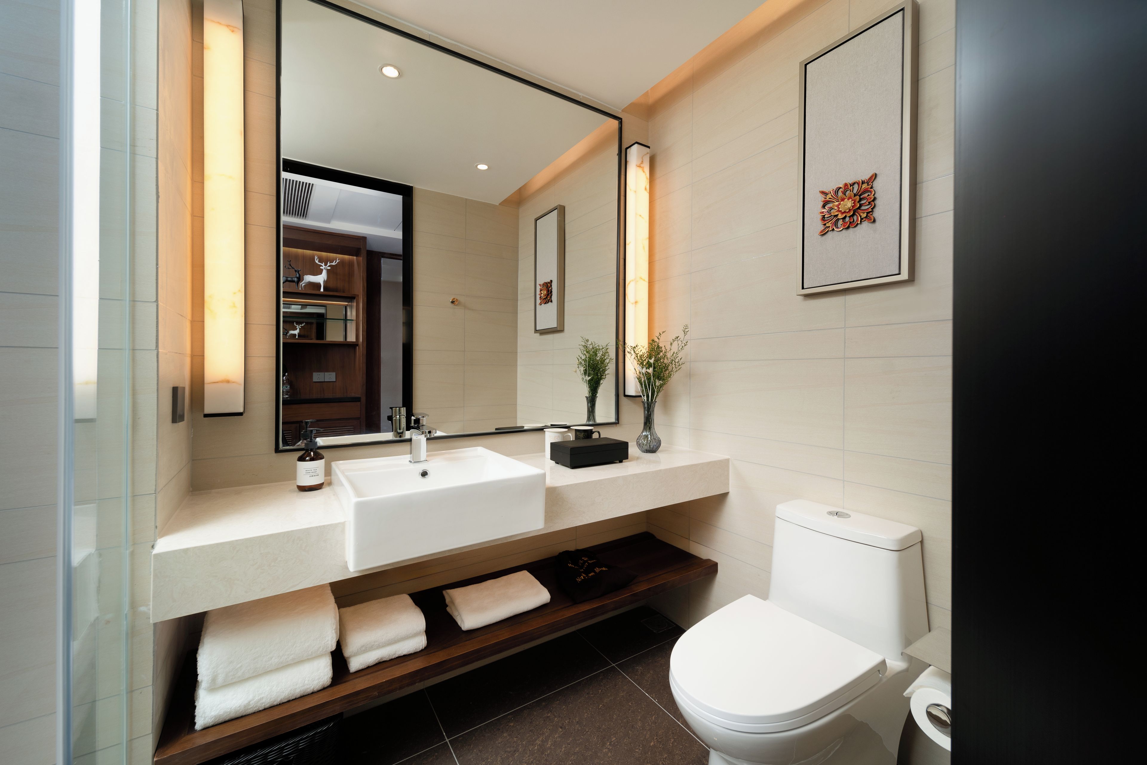 A modern bathroom with a large mirror, two sinks, a toilet, and a shelf with towels.