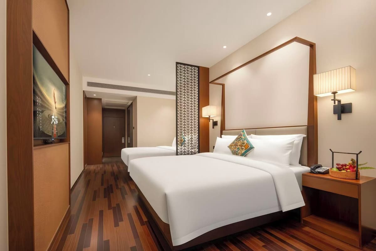 Habitación de hotel con dos camas, suelo de madera, panel decorativo en la pared, lámpara de noche y una mesita con frutas.