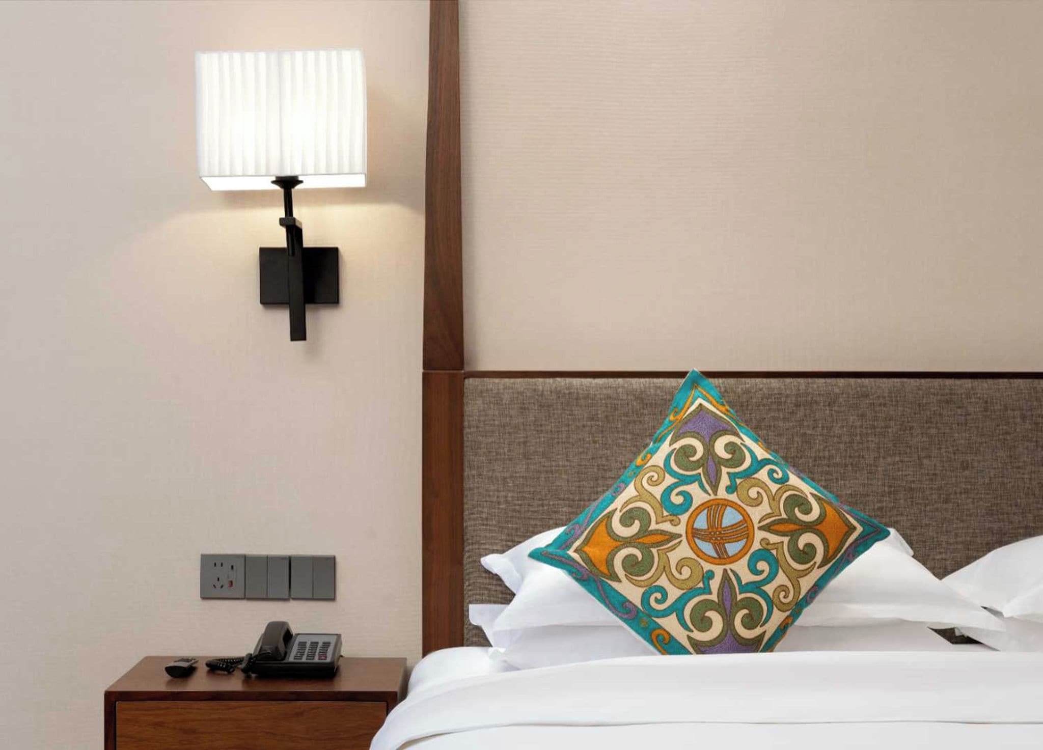 A hotel room with a bed, a nightstand with a telephone, a wall-mounted lamp, and a power outlet.