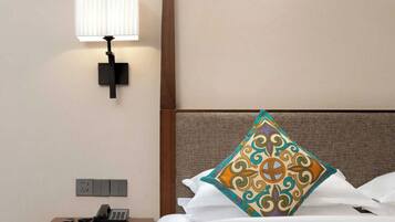 A hotel room with a bed, a nightstand with a telephone, a wall-mounted lamp, and a power outlet.