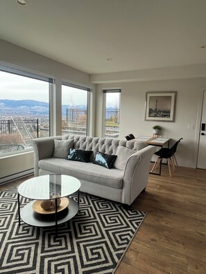 Ein modernes Wohnzimmer mit einer Couch, einem Glastisch, einem gemusterten Teppich, einem Essbereich mit Tisch und Stühlen sowie großen Fenstern mit Blick auf die Stadt.