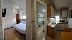 Une cuisine compacte avec un plan de travail en bois, un évier et un four à micro-ondes. La pièce adjacente contient un lit recouvert d’une couvre-lit à motifs.