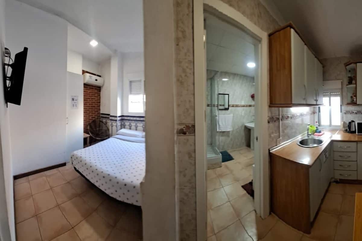 A compact kitchen with a wooden countertop, a sink, and a microwave. There is a bed with a patterned bedspread in the adjacent room.