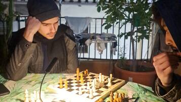 Two people playing chess at an outdoor table with a green tablecloth, surrounded by potted plants and a metal railing.