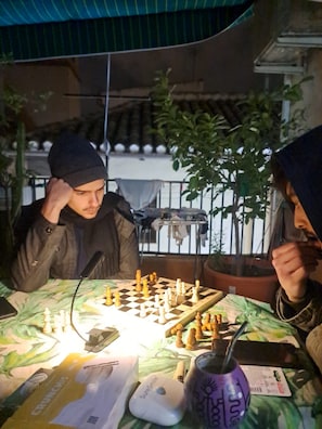 Two people playing chess at an outdoor table with a green tablecloth, surrounded by potted plants and a metal railing. - Hostal Cascabel (Granada)