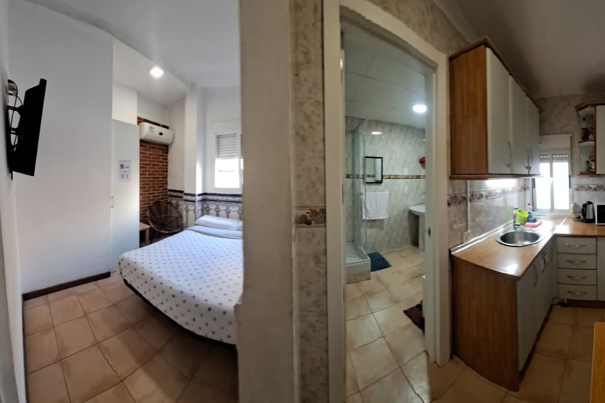 A compact kitchen with a wooden countertop, a sink, and a microwave. There is a bed with a patterned bedspread in the adjacent room.