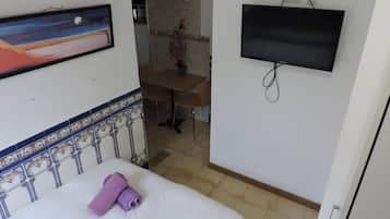 Quarto de hotel com uma cama, televisão fixada na parede, um quadro na parede e um pequeno espaço de refeições com uma mesa e cadeiras.