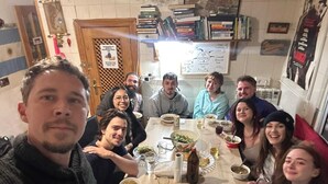 Un groupe de personnes est assis autour d’une table, devant différents plats, dans une pièce dotée d’une porte en bois et d’un tableau blanc mural.