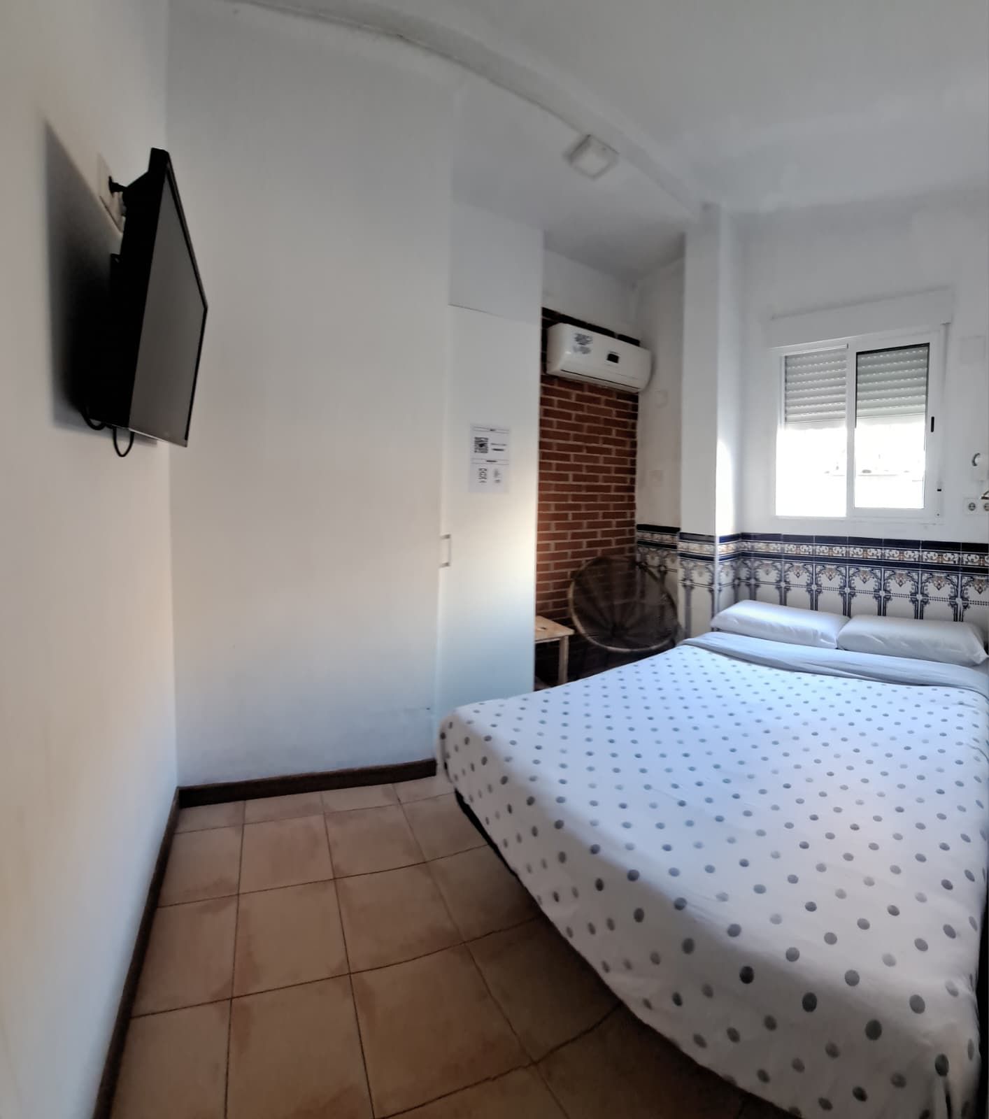 Quarto com cama, televisão na parede, janela com persianas e ar condicionado.