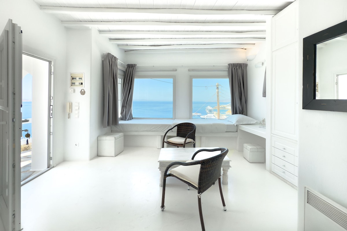 Habitació d'interior blanc, finestra amb vista al mar, taula blanca i cadira de vímet.
