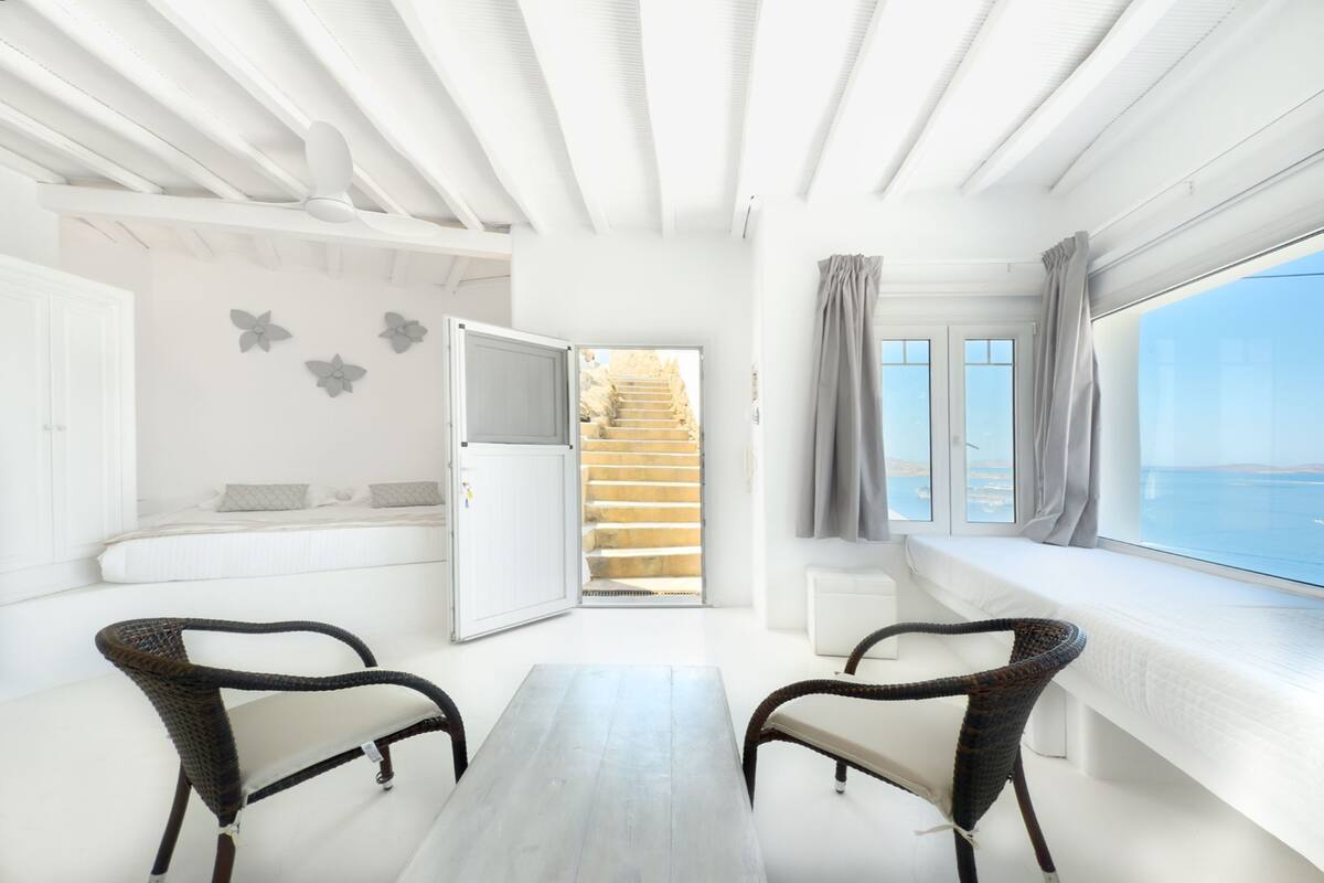 Een kamer met een wit plafond, een tafel, twee rieten stoelen, een bank, een raam met gordijnen en uitzicht op zee.