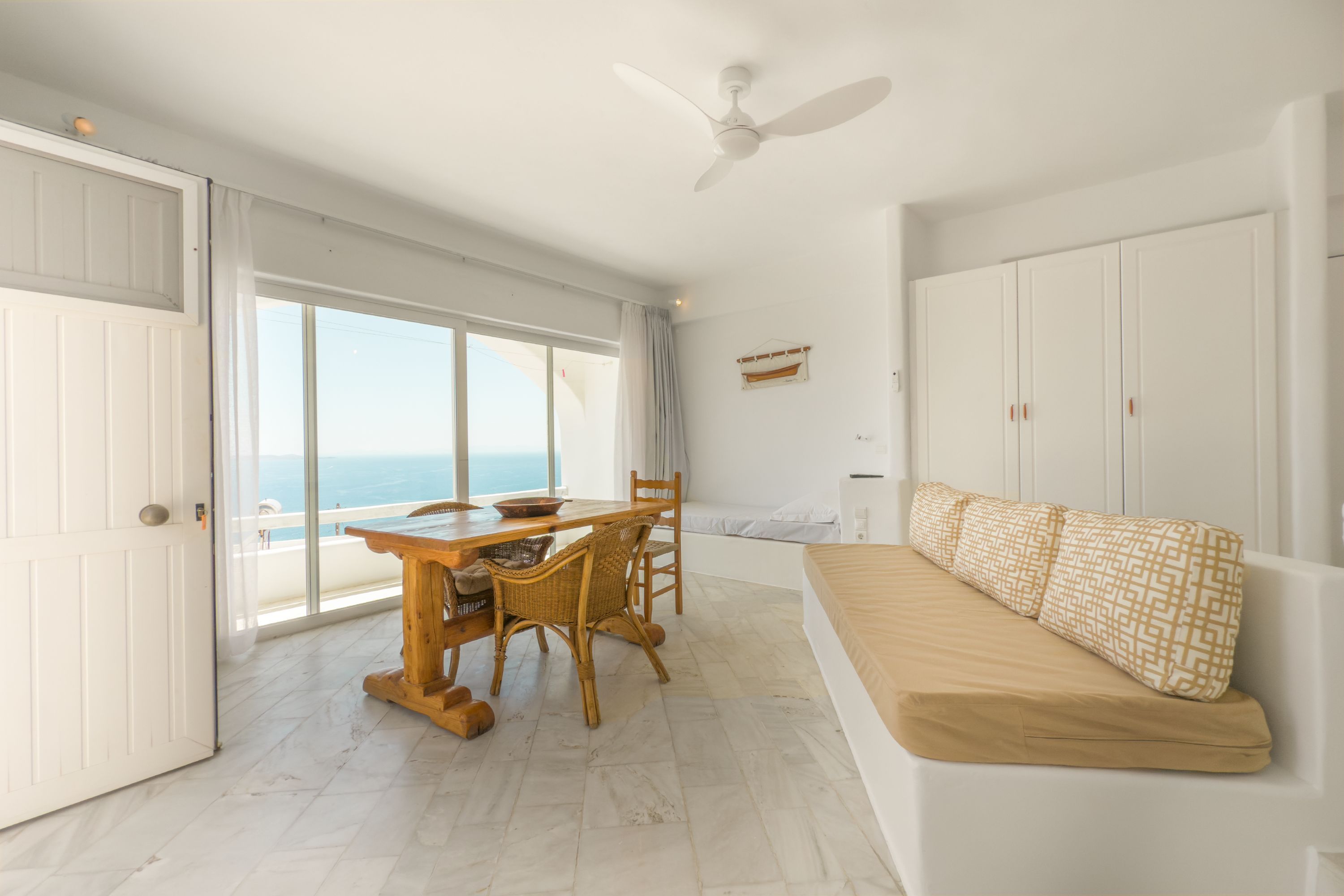 A room with a dining table, chairs, a sofa, a ceiling fan, and a view of the sea.