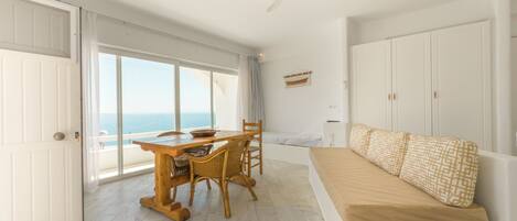 A room with a dining table, chairs, a sofa, a ceiling fan, and a view of the sea.