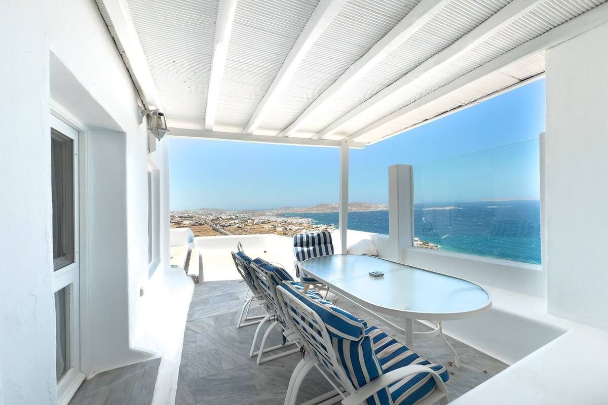 Een terras met tafel en stoelen, uitzicht op zee, en een wit interieur.