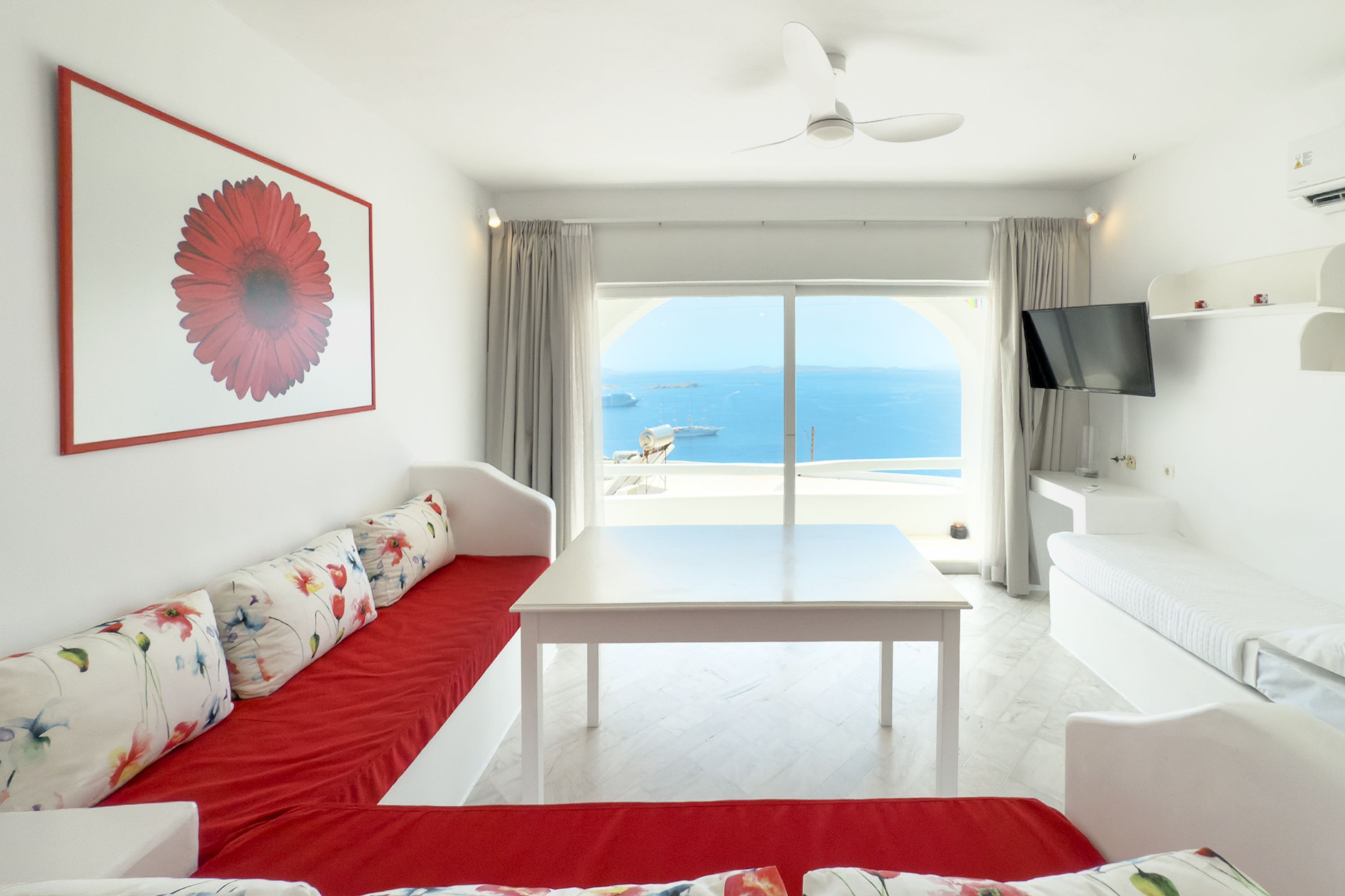 A modern room with a white sofa, a red accent, a table, a TV, and a large window with a view of the sea.