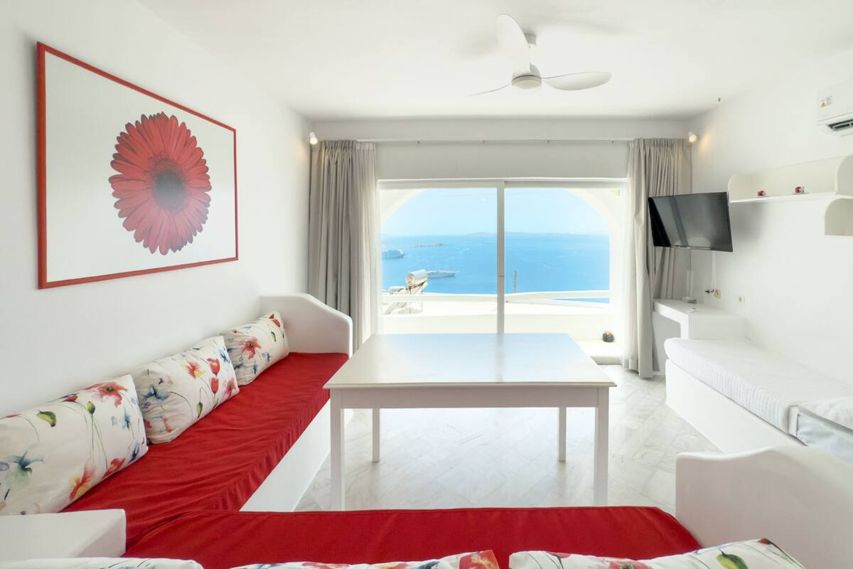 Een moderne kamer met een witte bank, een rode accentkleur, een tafel, een televisie en een groot raam met uitzicht op zee.