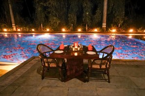 A dining table set for two with chairs, surrounded by a pool with floating rose petals and lights.