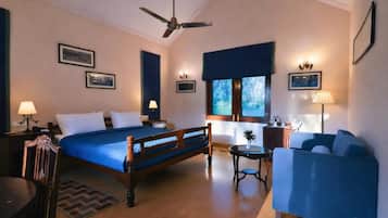 A hotel room with a bed, a desk, a chair, a sofa, a ceiling fan, and a window with a view of trees.