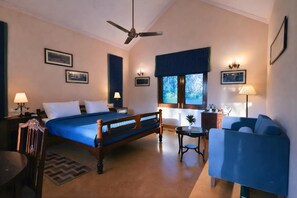 Ein Hotelzimmer mit Bett, Schreibtisch, Sessel, Sofa, Deckenventilator und Fenster mit Blick auf Bäume.