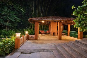 A covered outdoor seating area with a tiled roof, surrounded by greenery and stone steps. - Justa The Bagh, Kanha (Baihar)