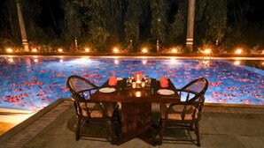 Une table à manger dressée pour deux personnes, avec des chaises, entourée d’une piscine où flottent des pétales de rose et où sont disposés des jeux de lumière.